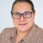 EIFA A person with long dark hair pulled back, wearing glasses and a beige, patterned blouse, smiles at the camera against a plain background, exuding the confidence of a bilingual professional.