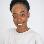 EIFA Une femme souriante aux cheveux courts et bouclés et aux boucles doreilles créoles pose dans un sweat-shirt de couleur claire avec un texte rouge, associé à un bandeau noir. Le fond blanc épuré renforce la simplicité de limage. Elle incarne lesprit dynamique de Londres, rappelant une journée joyeuse à lécole EIFA.