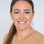 EIFA A bilingual woman with long brown hair tied back smiles at the camera. She is wearing a tan sweater and small gold earrings, embodying the charm of London style. The background is a plain light color.