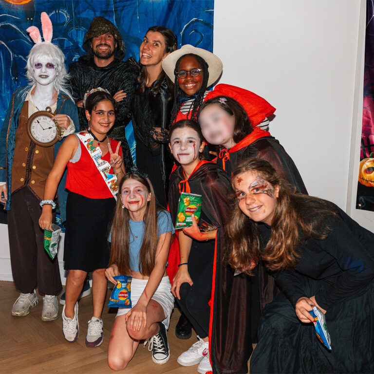 EIFA A bilingual group in Halloween costumes, featuring a rabbit-themed character with a clock, a hooded figure, and several others in capes and face paint. They pose indoors holding snacks at the EIFA event in London.