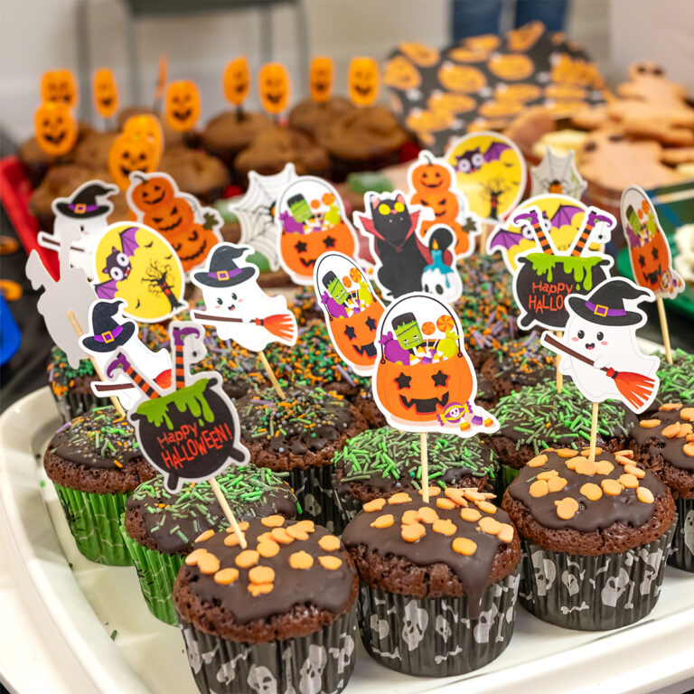 EIFA A delightful array of Halloween-themed cupcakes awaits at EIFAs London school event, topped with chocolate icing, sprinkles, and decorative picks featuring ghosts, pumpkins, and witches. Cookie faces and pumpkin treats in the background add to the festive spread.