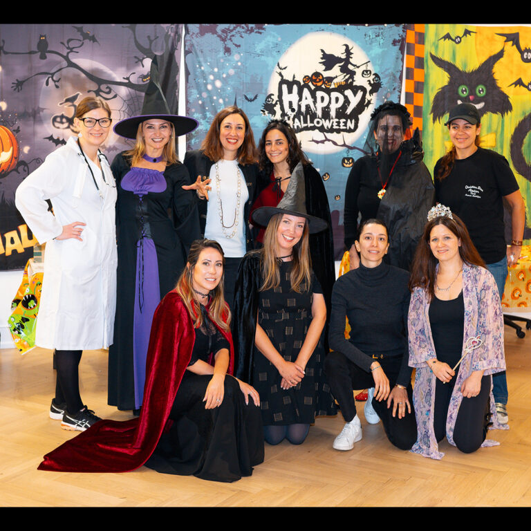 EIFA A bilingual group of people posing for a photo in Halloween costumes, including witches, a princess, and a doctor. They are smiling and standing in front of festive Halloween decorations like pumpkins and banners at an EIFA event in London.
