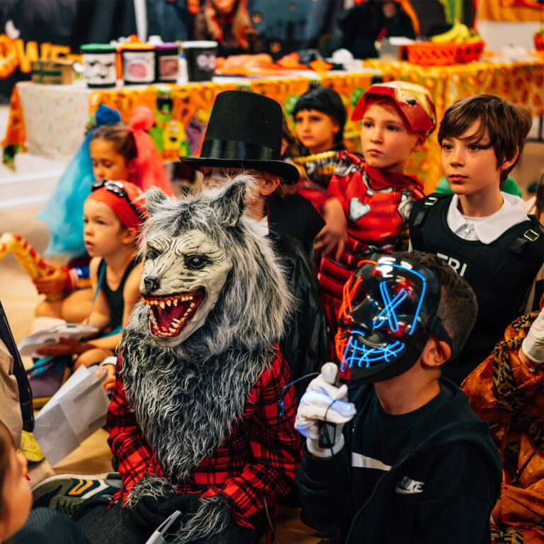 EIFA A group of children in colorful Halloween costumes sit together indoors at the EIFA school in London. Outfits include a werewolf, superhero, and a masked figure with glowing lines. The bilingual setting enriches the festive, playful atmosphere with decorations all around.