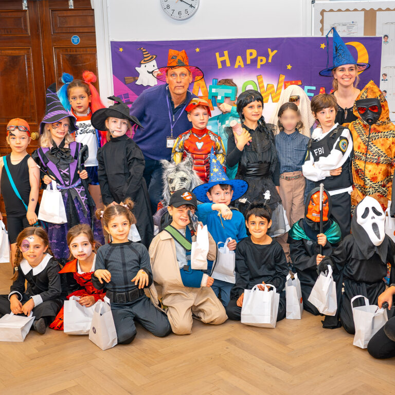 EIFA A group of children from EIFA, dressed in various Halloween costumes, pose for a photo with two adults in front of a purple Happy Halloween banner. The kids hold white bags and are dressed as witches, superheroes, and other spooky characters on the wooden floor of their bilingual school.
