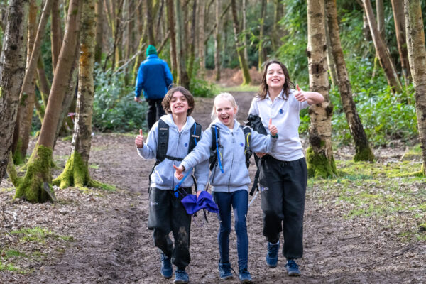 EIFA Three children, wearing backpacks and casual outdoor clothing, from a bilingual school joyfully walk on a forest trail, giving thumbs up. Bare trees line the path, and a person in a blue jacket walks ahead. The ground is muddy, suggesting a recent rain.