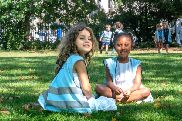 EIFA Two young children sit on the grass in their London school uniforms, wearing light blue and white outfits with reflective stripes. One has curly hair and the other a bun. They smile, surrounded by trees and other EIFA students dressed in similar attire in the background.