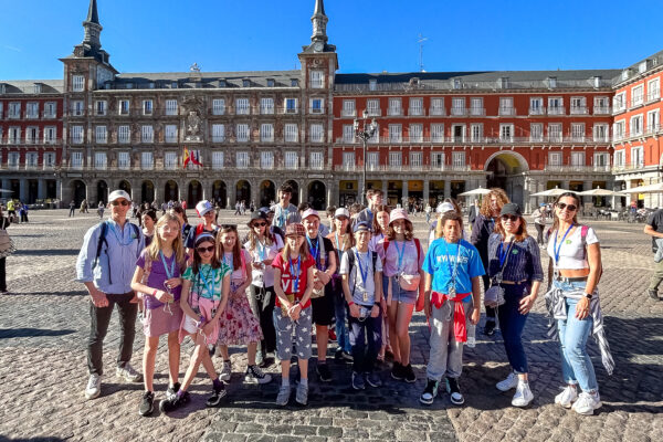 EIFA A group of people, mostly wearing casual clothes and sunglasses, stand in a sunny cobblestone plaza outside a historic building in London. The scene captures the relaxed atmosphere near the esteemed EIFA International School under a clear, blue sky.