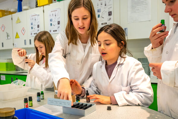 EIFA Four bilingual children in lab coats engage in a science experiment at a London school laboratory table. They handle small bottles and a box, appearing focused and collaborative. The background showcases lab equipment and safety posters, fostering an environment of learning and discovery.