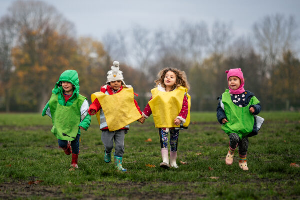 EIFA Four children from a bilingual school in London joyfully run across a field, wearing colorful winter clothing and vibrant yellow and green vests. The background shows a cloudy sky and trees with autumn leaves.