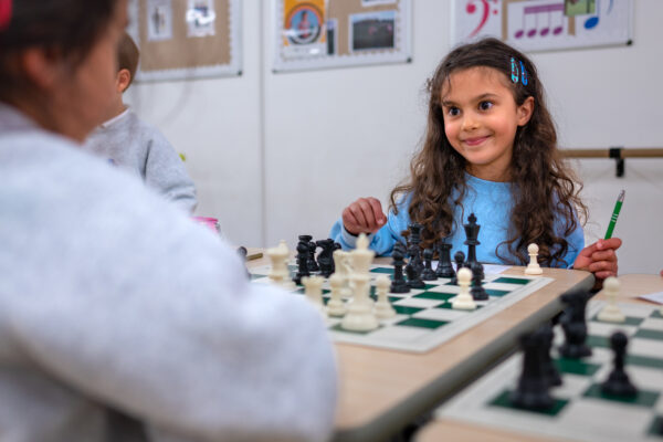 EIFA A young girl with long curly hair, wearing a blue sweater and holding a pen, smiles as she plays chess with an opponent. The chessboard is in focus, set against the backdrop of a vibrant classroom in a school in London.