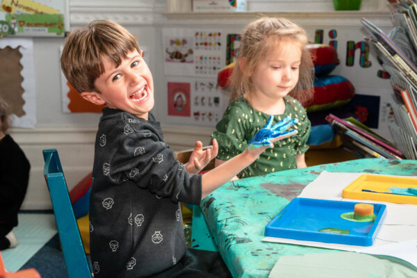 EIFA In a vibrant London classroom, two children are seated at a table draped with a green tablecloth. The boy on the left, smiling proudly, displays his paint-covered hand, while the girl on the right focuses intently on her artwork. Its a joyous scene in this bilingual school setting.