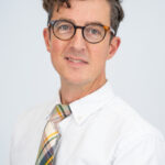 EIFA A person with short, wavy brown hair and glasses is wearing a white shirt and a colorful plaid tie, embodying the essence of EIFAs bilingual school spirit. They face the camera with a neutral expression against a plain background.