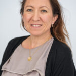 EIFA A bilingual woman with long brown hair is wearing a light gray blouse and a black cardigan. She smiles slightly at the camera, her simple necklace resting on her neck. The plain, light-colored background hints at a sophisticated setting in London.
