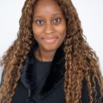 EIFA A bilingual woman with long, wavy brown hair smiles gently at the camera. She is wearing a black top with a textured material and a small bow near the neckline. The plain white background gives the image a clean, professional feel reminiscent of a school portrait in London.