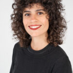 EIFA A person with curly brown hair smiles at the camera, wearing a black shirt, embodying the vibrant spirit of London against a plain white background.