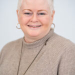EIFA A person with short, white hair smiles at the camera. They are wearing a beige sweater and hoop earrings against a plain, light-colored background, exuding the warmth of a bilingual teacher from EIFA School.