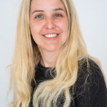 EIFA A person with long blonde hair, wearing a black top, smiles warmly at the camera. The plain, light-colored wall behind them hints at a serene environment. Perhaps theyre capturing a moment outside their London school, where days are filled with learning and laughter at EIFA.