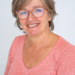 EIFA A smiling bilingual woman with short gray hair wears a pink long-sleeve shirt and glasses, set against a light background.