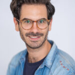 EIFA A bilingual man with brown hair and beard, wearing glasses, a denim jacket, and a dark shirt, smiles slightly while facing forward against a plain white background. His style hints at a scholarly vibe that wouldnt be out of place in the creative streets of London.