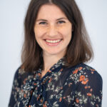 EIFA A woman with shoulder-length brown hair smiles warmly. She is wearing a dark floral blouse with a bow at the neck. The background is a soft gray, creating a neutral setting that highlights her friendly expression, reminiscent of her bilingual experiences in London.
