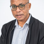 EIFA A bilingual man wearing glasses, a black jacket, and a striped shirt stands against a light backdrop. His bald head and goatee add to his neutral expression as he gazes at the camera, embodying the diverse spirit of London.