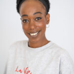 EIFA A person smiling at the camera, wearing a light gray sweatshirt with red text saying La Vie en couleur! They have short curly hair, a nose ring, hoop earrings, and a black headband. With a style as vibrant as London itself, they embrace the bilingual flair represented at EIFA. The background is plain white.