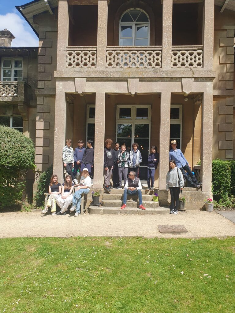 EIFA Un groupe de personnes, certaines assises, dautres debout, posent sur les marches dun grand bâtiment historique en pierre de Londres. Lentrée est surmontée dun balcon. La verdure et une pelouse ensoleillée entourent la scène, ajoutant du charme à ce rassemblement bilingue à lEIFA.