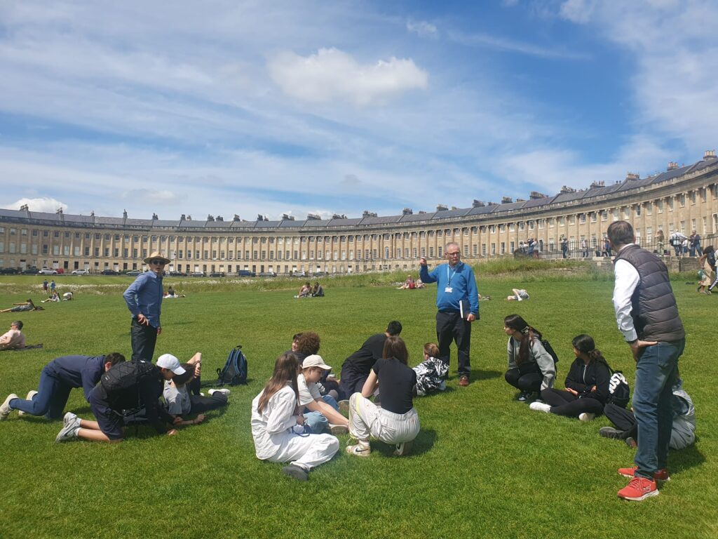 EIFA Un groupe de personnes est assis sur lherbe avec un guide bilingue qui discute avec eux. Ils se trouvent devant le Royal Crescent, un monument géorgien emblématique qui rappelle le charme architectural de Londres, sous un ciel partiellement nuageux. Certaines personnes sont allongées et profitent de la journée ensoleillée.