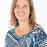 EIFA A person with shoulder-length brown hair smiles at the camera, wearing a blue and white paisley-patterned top and a necklace. The portrait exudes a sense of academic elegance, reminiscent of an inspiring day spent at an EIFA school in London, set against a plain white background.