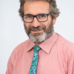 EIFA A man with glasses and a graying beard smiles while wearing a pink shirt and a blue-green floral tie. The background is plain light gray, emphasizing a professional appearance, as if prepared for an event at the prestigious EIFA School in London.