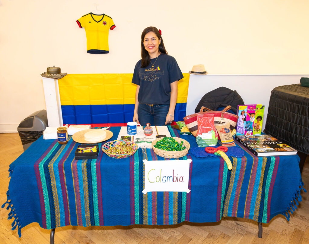 EIFA A woman stands behind a table at the EIFA school event, displaying Colombian cultural items. Theres a blue tablecloth with food, books, and crafts. A Colombian flag and jersey hang in the background, embodying bilingual pride. A sign on the table reads Colombia.