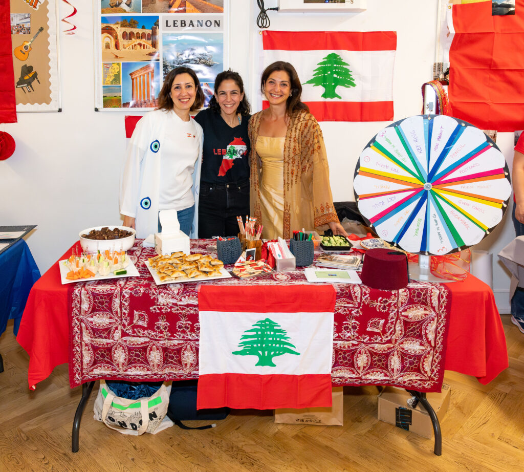 EIFA Three people stand smiling behind a table decorated with Lebanese flags and foods at the EIFA event in London. Behind them is a display of Lebanese images and a colorful prize wheel. The bilingual exhibit features traditional attire and snacks, celebrating rich cultural heritage.
