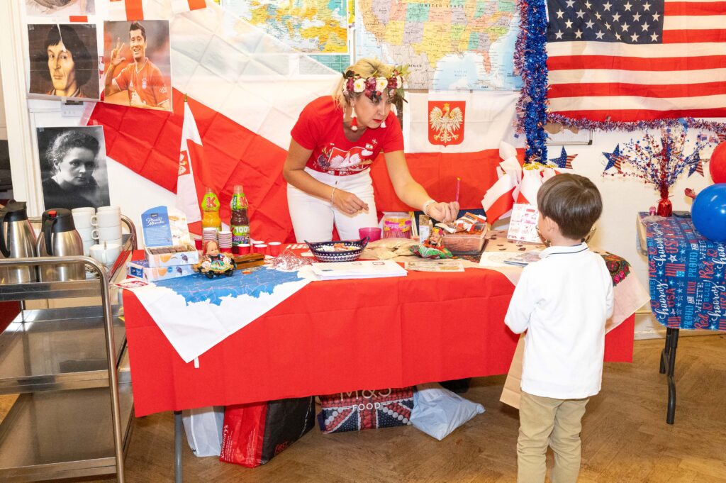 EIFA A woman in a red shirt and white headband stands at a table covered with Polish flags, souvenirs, and decorations, engaging with a young boy in a white shirt. An American flag hangs in the background of this bilingual school event in London.
