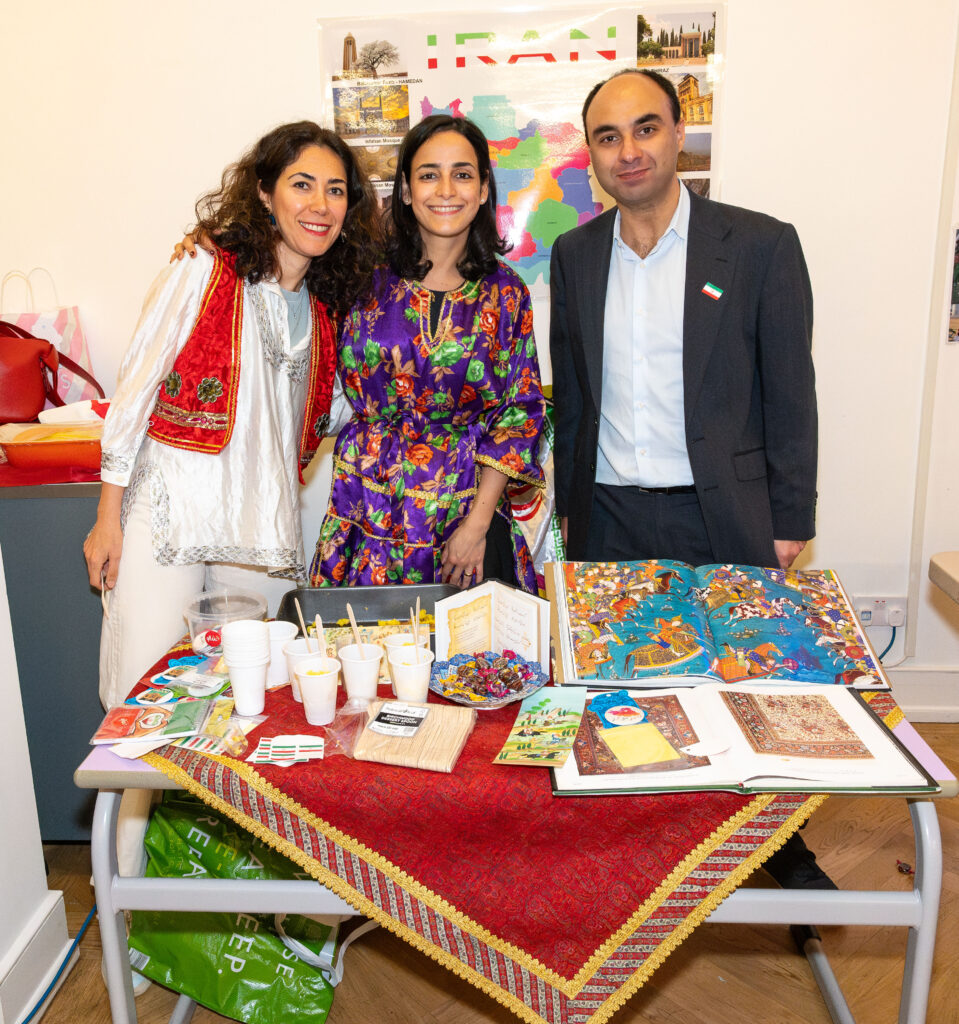 EIFA Three people stand behind a table at the EIFA school in London, displaying cultural items, books, and snacks. They are smiling, wearing colorful traditional clothing, with a map of Iran in the background. The atmosphere is vibrant and engaging.
