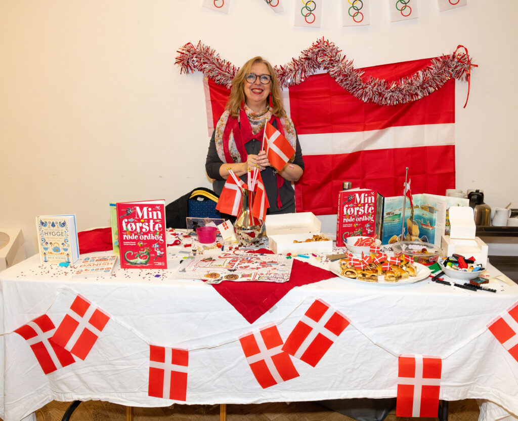 EIFA A woman stands behind a table decorated with Danish flags and assorted items at the bilingual school fair. Books, snacks, and crafts are displayed. A large Danish flag and tinsel are hung on the wall behind her. She smiles, holding two small flags, representing EIFAs diverse community.
.