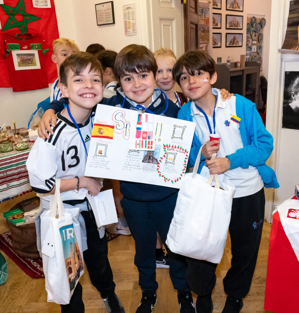 EIFA A group of smiling children from EIFA in London stand close together, holding a colorful poster with various international flags and the word Spain! written on it. These bilingual kids are wearing lanyards and holding white tote bags against a backdrop of vibrant cultural displays.