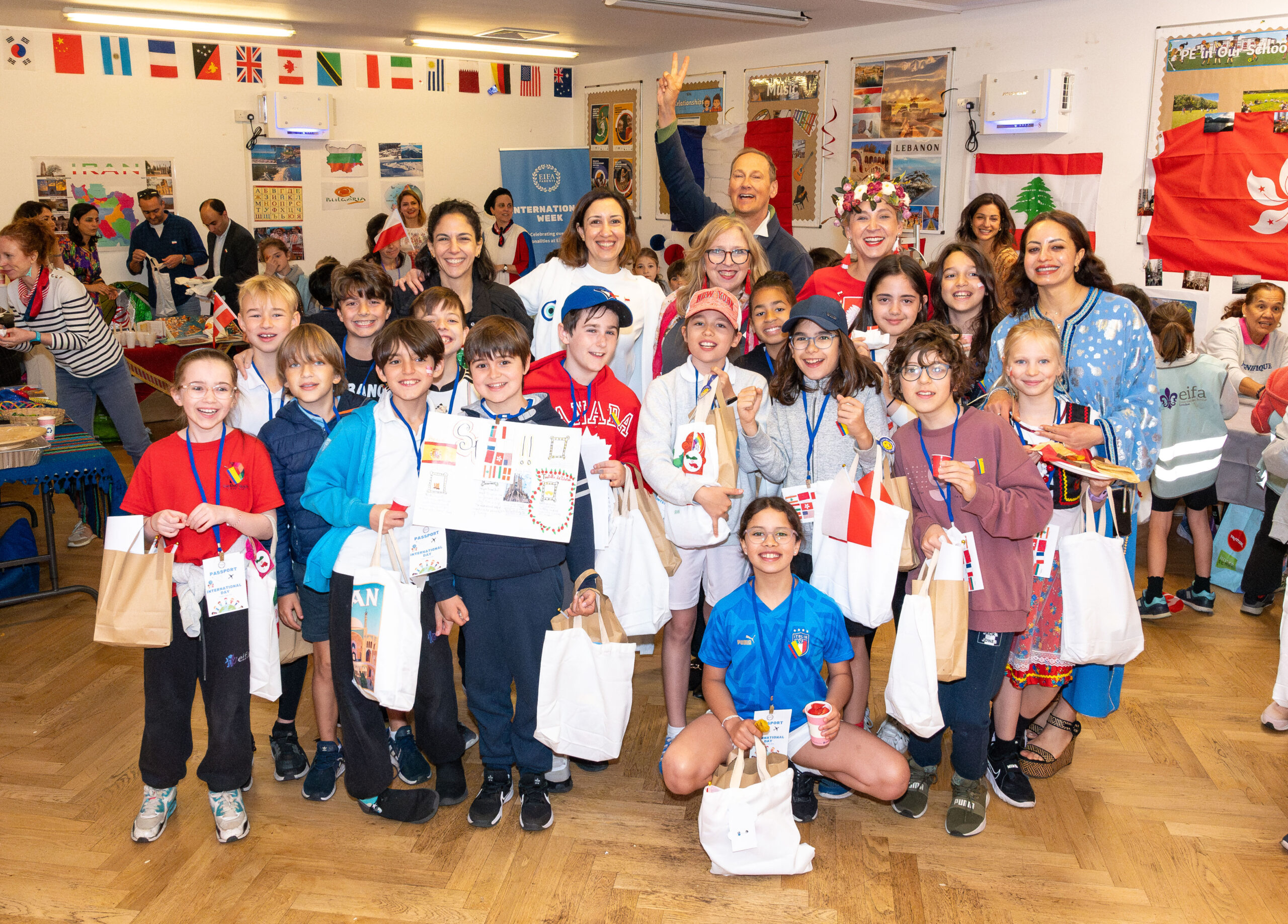 EIFA A diverse group of children and adults smiles in a colorful London classroom at EIFA School. The children hold paper crafts and gift bags, while international flags decorate the room, creating a festive atmosphere as everyone appears to be celebrating an event.