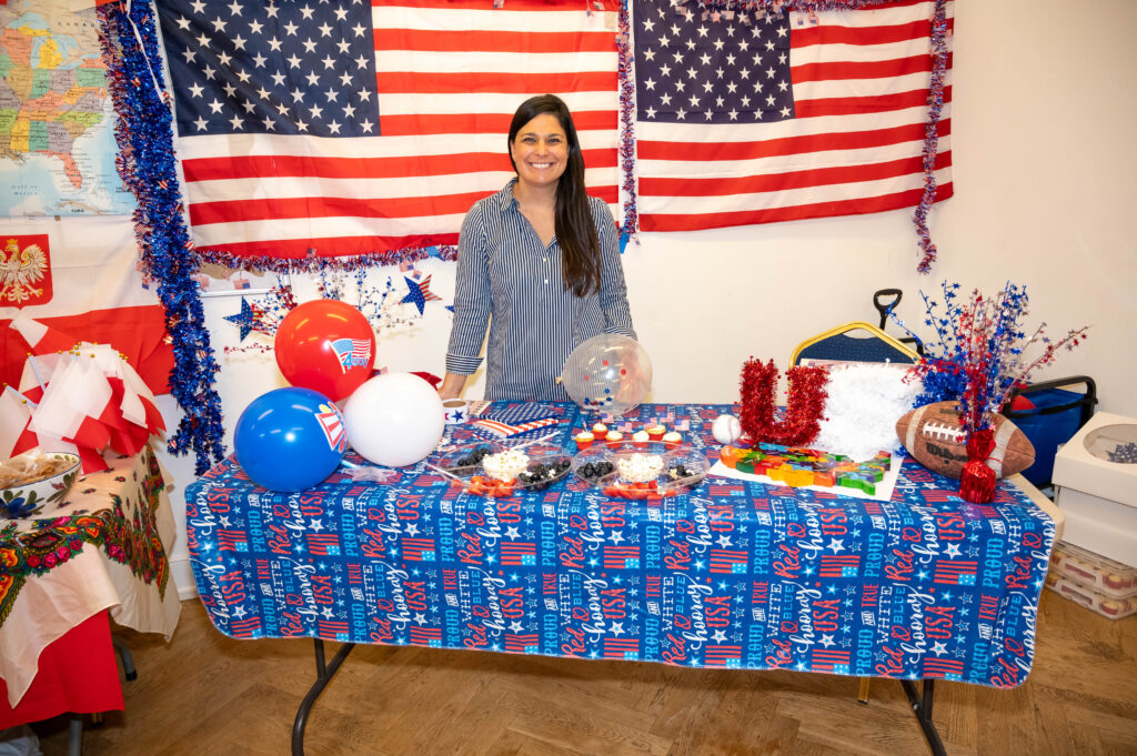 EIFA A person stands smiling behind a table with a red, white, and blue theme at the EIFA bilingual school event. The table has assorted items, balloons, and festive decorations. American flags hang in the background on the wall.