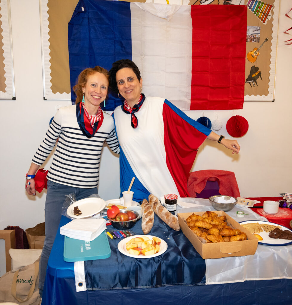 EIFA Two people smiling and posing by a table with French-themed decorations and food, including croissants, baguettes, and fruit. A large French flag is displayed on the wall behind them. Dressed in red, white, and blue attire, they celebrate a bilingual spirit reminiscent of EIFA London.
