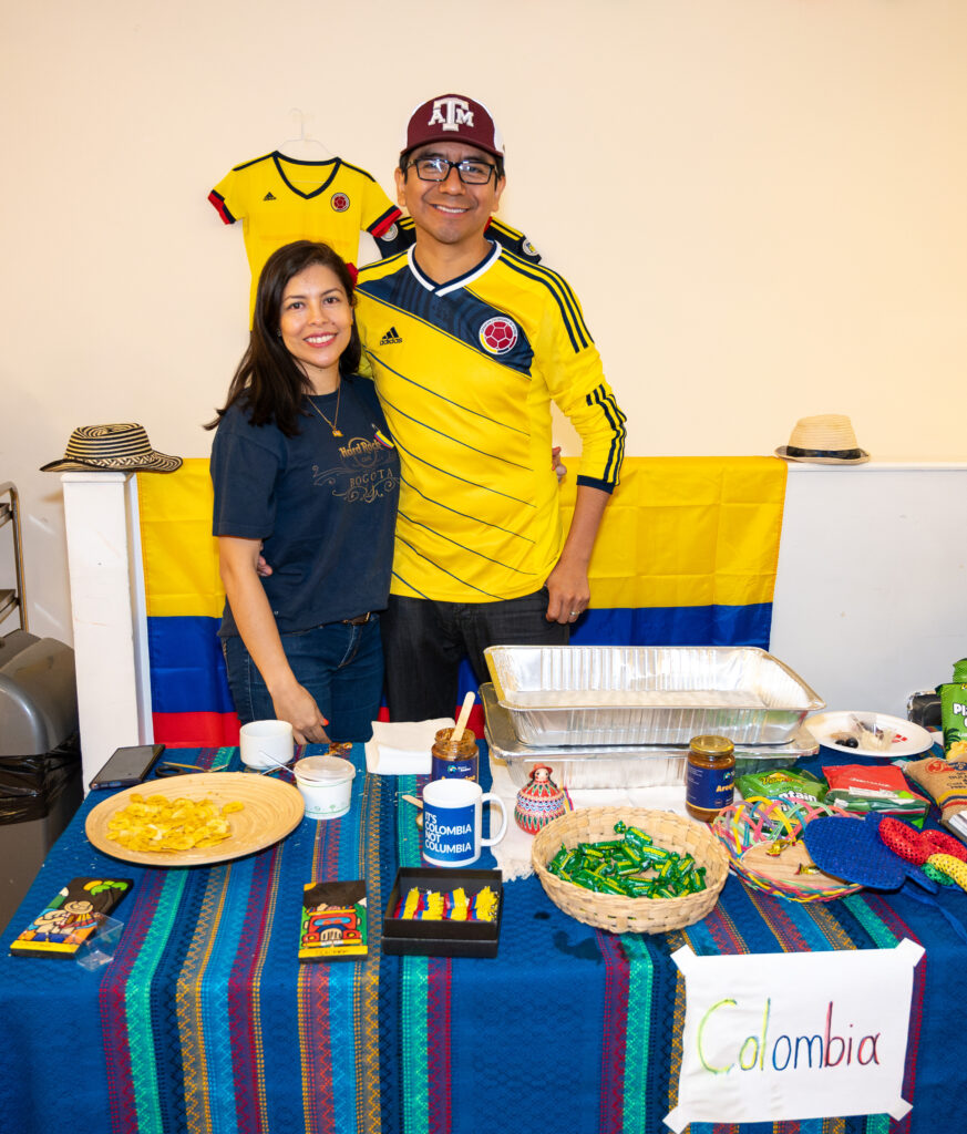EIFA A smiling couple stands by a table adorned with Colombian snacks and decorations, including flags and jerseys. The man sports a Colombian jersey and cap while the woman wears a navy shirt. A Colombia sign decorates the tablecloth, capturing the lively spirit at their London celebration.