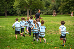 EIFA A group of children wearing blue and white vests, likely from a local school in London, run on the grass in a park, following an adult in a black shirt. Trees and greenery surround the open area, creating a lively and energetic atmosphere.