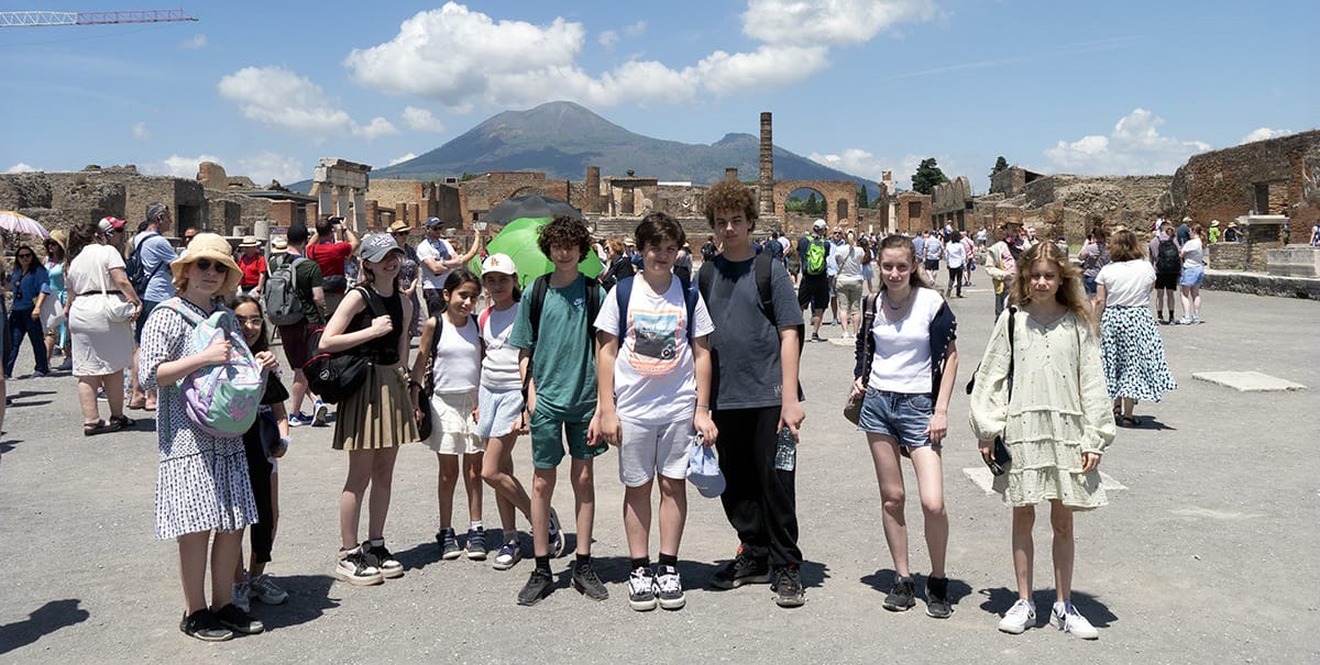 EIFA A group from EIFA School in London, including kids, stands in front of ancient ruins with Mount Vesuvius as the backdrop. Its a sunny day, and they are dressed in casual summer clothing. People and ruins complete this picturesque setting.