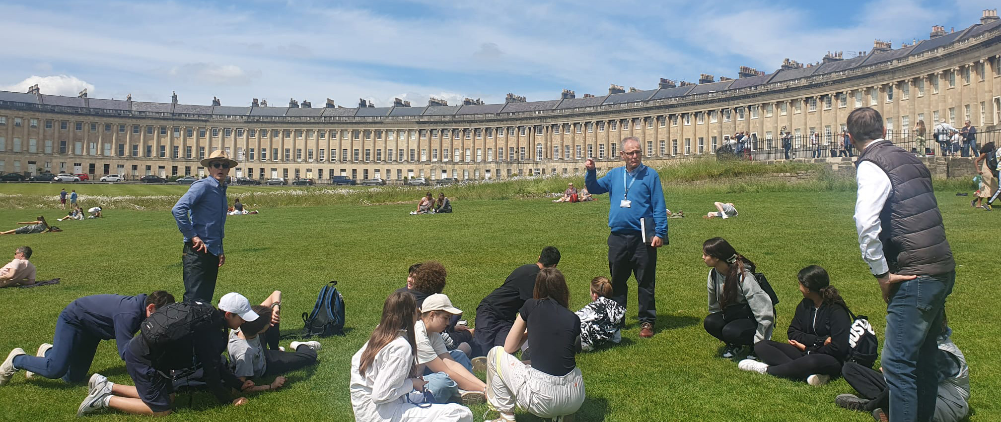 EIFA A group of people sit and stand on a grassy area in front of a historic, curved Georgian building under a blue London sky. A man in a blue sweater appears to be speaking, while others relax and listen. Some bilingual individuals sit or lie on the grass nearby.