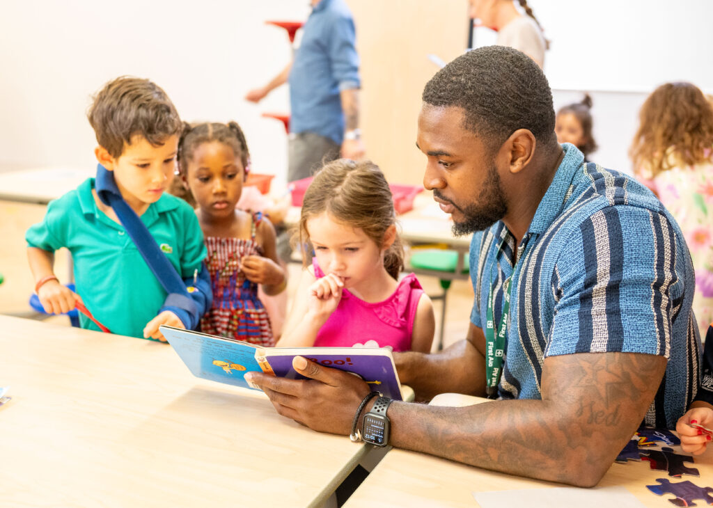 EIFA A man reads a colorful bilingual book to a group of three young children gathered around a table at EIFA. The children are intently looking at the book, captivated by the story. Other children and adults are visible in the background, creating a lively school atmosphere.