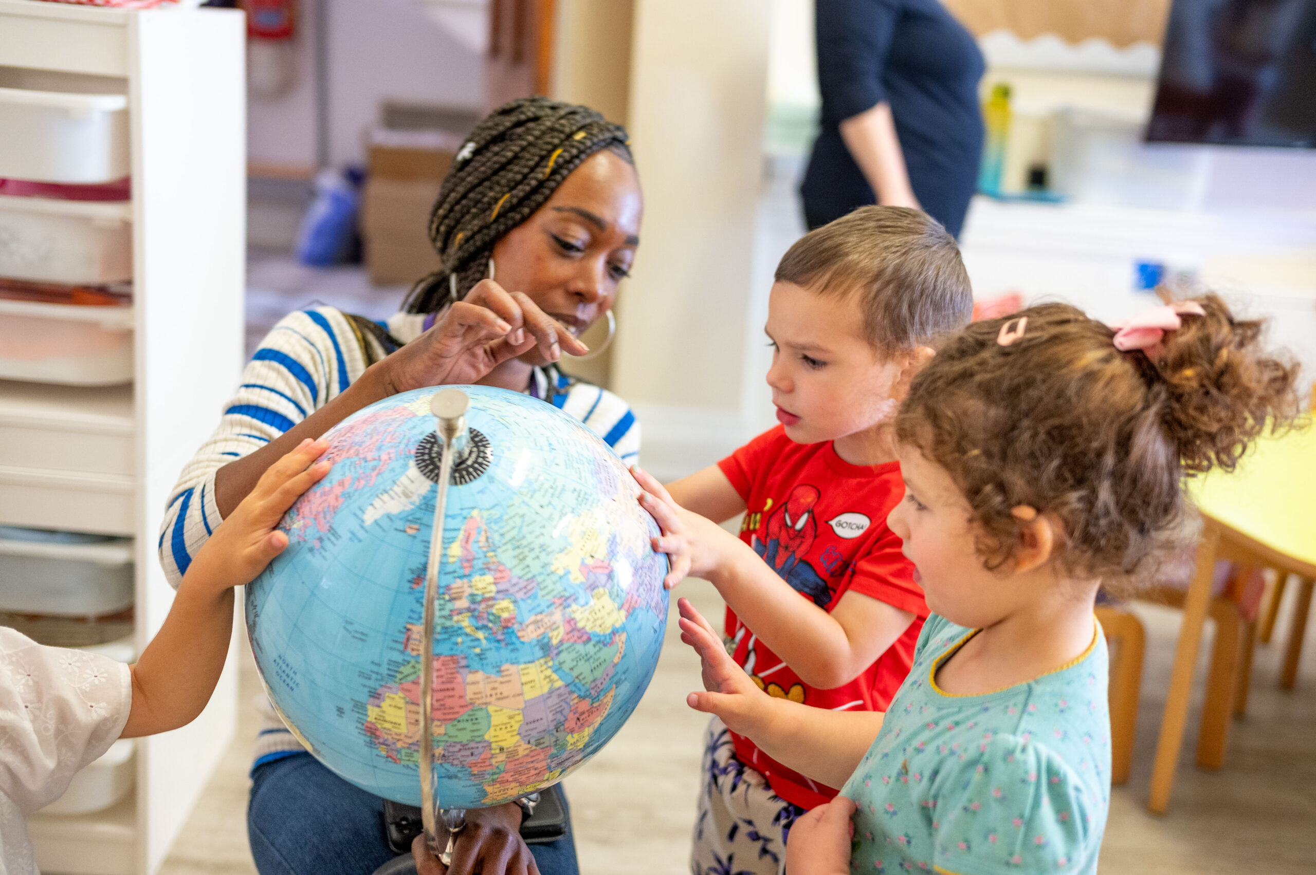 EIFA A woman and three young children are gathered around a globe in their bilingual school. The woman is pointing to a location on the globe, possibly London, while the children look on with interest. The setting appears to be a classroom with shelves in the background.