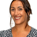 EIFA A woman with long dark hair smiles warmly. She is wearing a polka dot blouse and a delicate gold necklace. The plain white background subtly hints at an EIFA school environment, reflecting her bilingual nature.