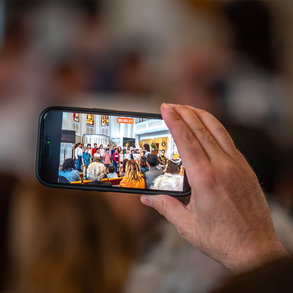 EIFA Une personne tient un smartphone et filme une chorale bilingue se produisant dans une église. La salle est dotée de bancs en bois, dun grand vitrail et de plusieurs personnes assises au premier plan.
