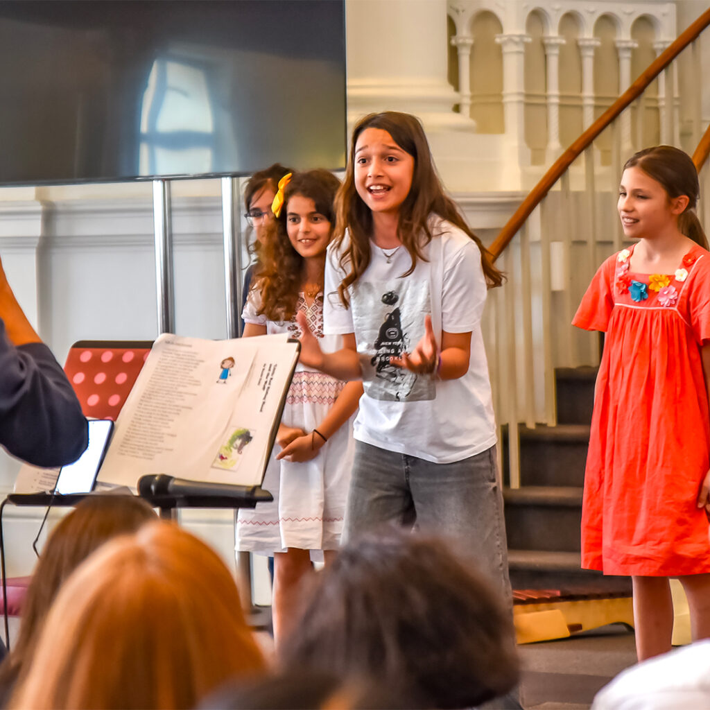 EIFA Trois filles se produisent avec enthousiasme sur scène dans une salle lumineuse avec un escalier incurvé, rappelant un grand auditorium décole à Londres. Une fille en chemise blanche fait des gestes passionnés au centre, flanquée de filles en robes blanches et orange. Un public bilingue est visible de derrière.
.