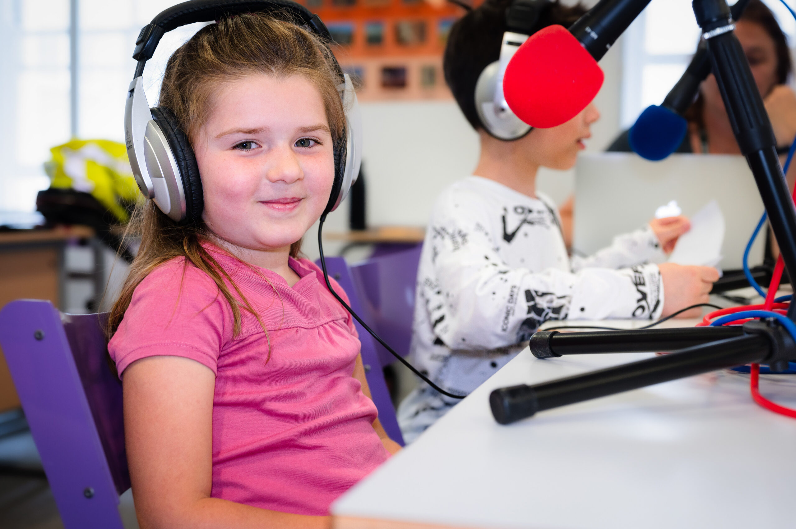 EIFA A young girl wearing headphones sits at a table with a microphone, smiling at the camera. A bilingual boy next to her speaks into another microphone. The background reveals school furniture and a bulletin board, suggesting theyre in a bustling London classroom.