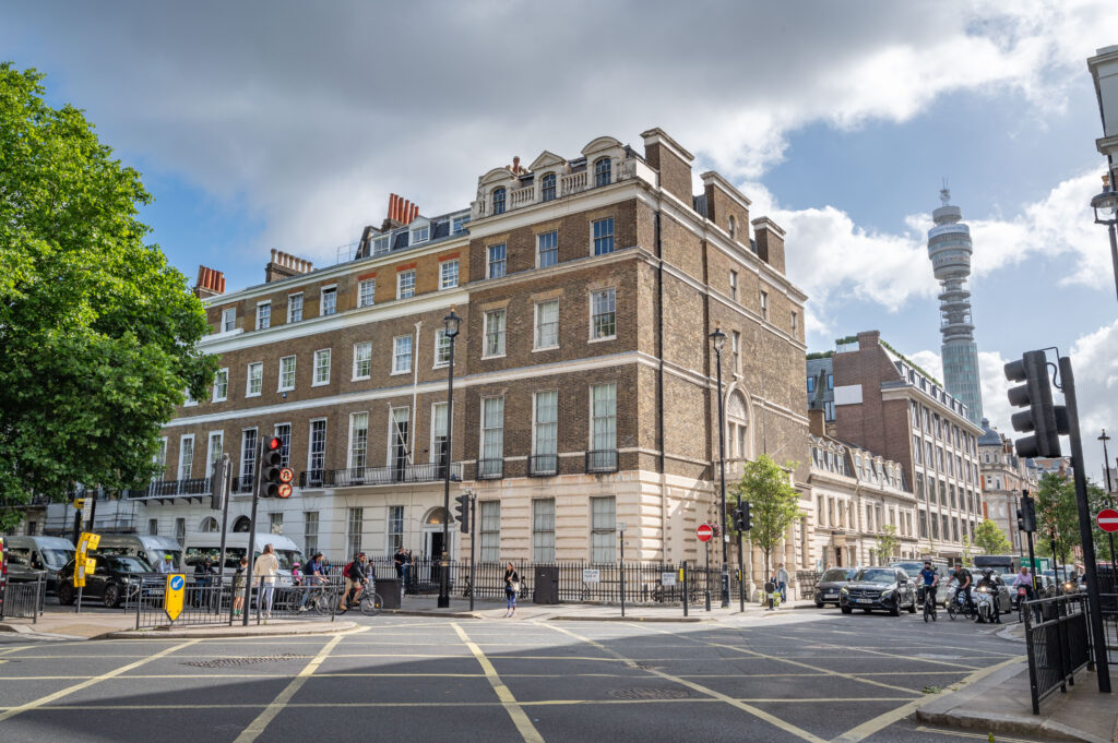 EIFA Une vue urbaine animée de Londres présente des bâtiments historiques en briques sous un ciel partiellement nuageux. Lemblématique BT Tower sélève en arrière-plan tandis que des habitants bilingues se promènent sur les trottoirs, tandis que les voitures sarrêtent à lintersection sous les feux de circulation.
